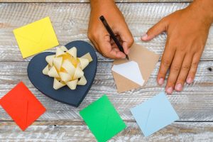 birthday-concept-with-gift-box-envelopes-wooden-background-flat-lay-man-writing-greeting-card-with-pen_176474-7605-1