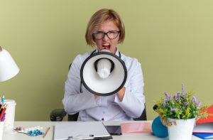 angry-young-female-doctor-wearing-medical-robe-with-glasses-stethoscope-sits-table-with-medical-tools-speaks-loudspeaker-isolated-olive-green-wall_141793-105622