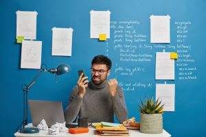 annoyed-male-boss-works-home-cozy-workplace-organizes-working-process-clenches-fist-teeth-with-irritation_273609-34349