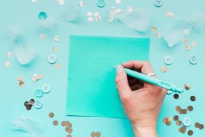close-up-woman-s-hand-writing-paper-with-pen-surrounded-with-feather-sequins-button-teal-backdrop_23-2148125298