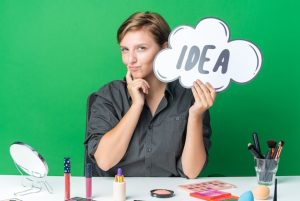impressed-beautiful-woman-sits-table-with-makeup-tools-holding-idea-bubble-with-makeup-brush-putting-finger-cheek_141793-121662