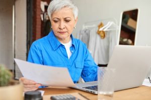 people-lifestyle-domesticity-modern-technology-concept-concentrated-retired-woman-with-short-gray-hair-holding-sheet-paper-doing-domestic-finances-home-using-laptop-calculator_343059-2821