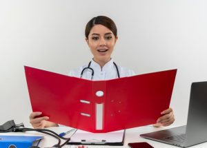 pleased-young-female-doctor-wearing-medical-robe-with-stethoscope-sitting-desk-work-computer-with-medical-tools-holding-looking-folder-with-copy-space_141793-37717
