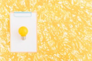 yellow-light-bulb-white-paper-clipboard-against-yellow-backdrop_23-2147873434