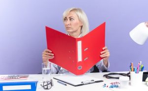 confident-adult-slavic-female-doctor-medical-robe-with-stethoscope-sitting-desk-with-office-tools-holding-file-folder-looking-side-isolated-purple-background-with-copy-space_141793-105223