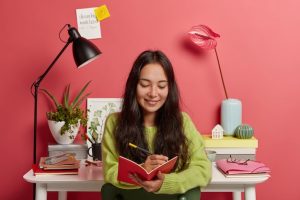glad-concentrated-woman-focused-notepad-writes-down-ideas-essay-research-work-makes-up-review-poses-against-workplace-with-desk-lamp_273609-34061